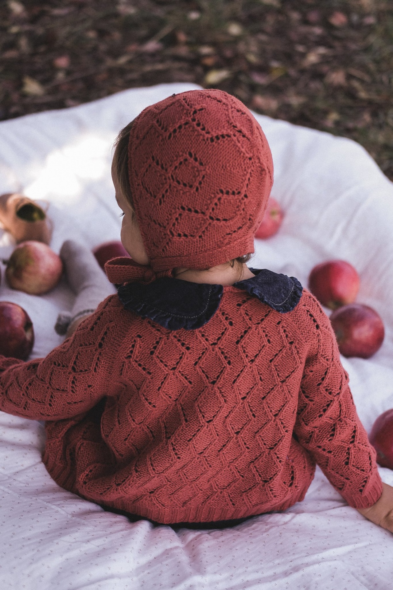 Criança com roupa de malha vermelha e gorro sentada com maçãs ao redor