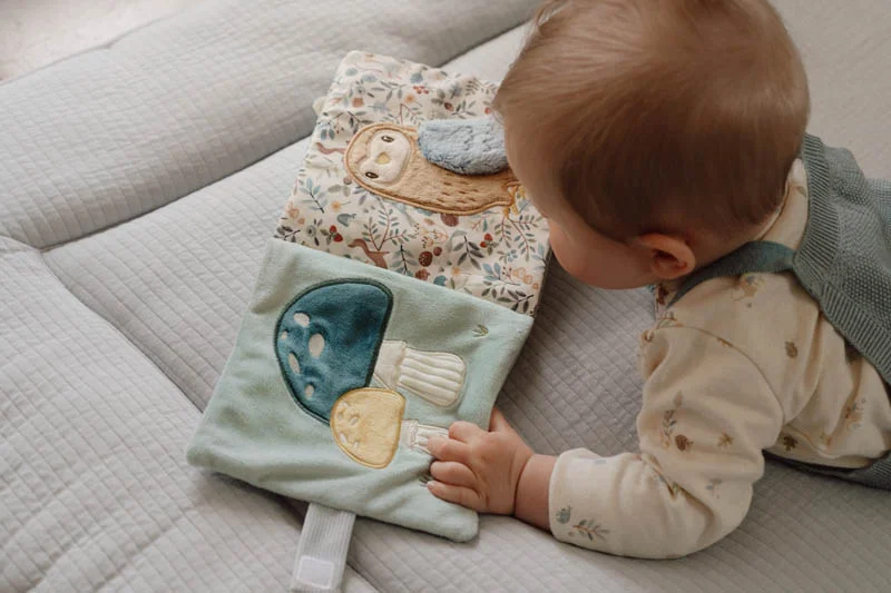 Livro macio de tecido para bebés com ilustrações bordadas de cogumelos e coruja ao lado de bebé a tocar o livro