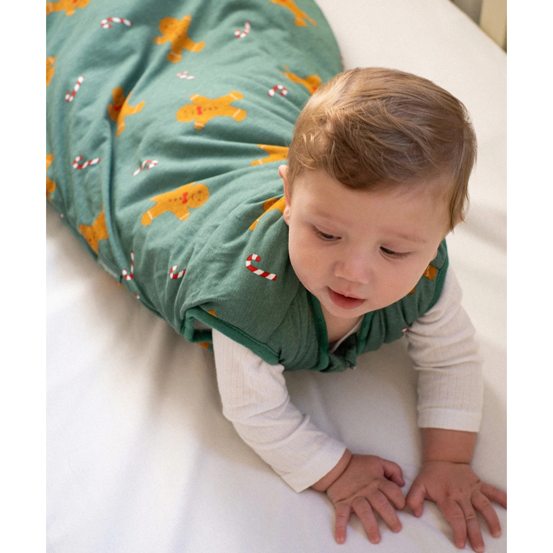 Bebé com saco-cama verde com padrão de bonecos de gengibre e bastões de açúcar a rastejar sobre cama branca