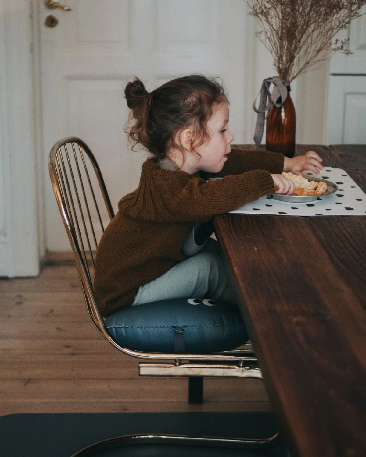 Criança sentada numa cadeira metálica com almofada azul, à mesa de madeira com manta branca com pontos pretos, vaso castanho com plantas secas ao fundo.