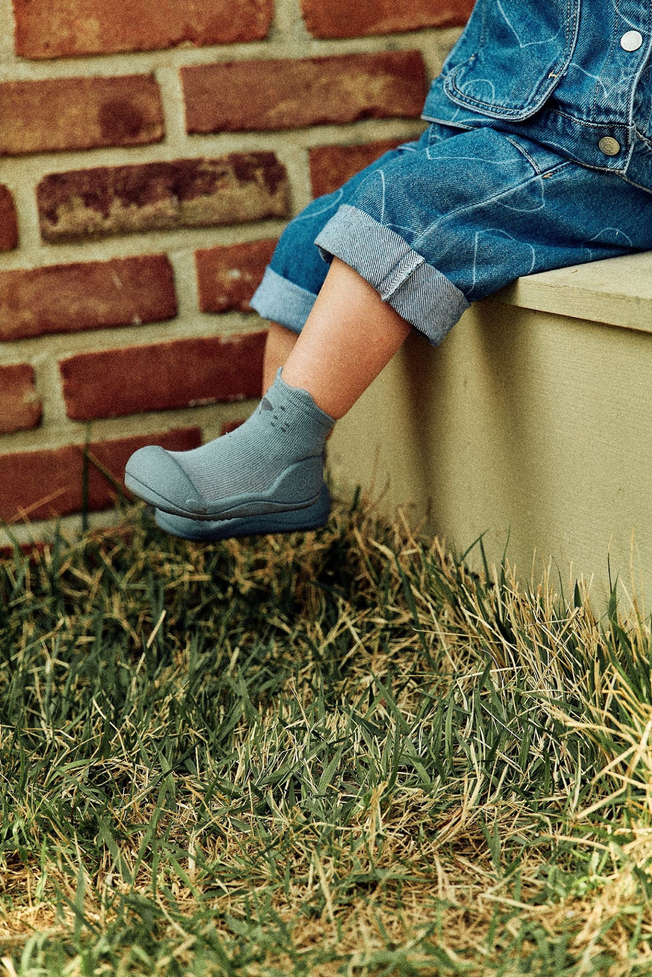 Sapatos infantis cinzentos usados com calças de ganga azuis junto a muro de tijolos e relva