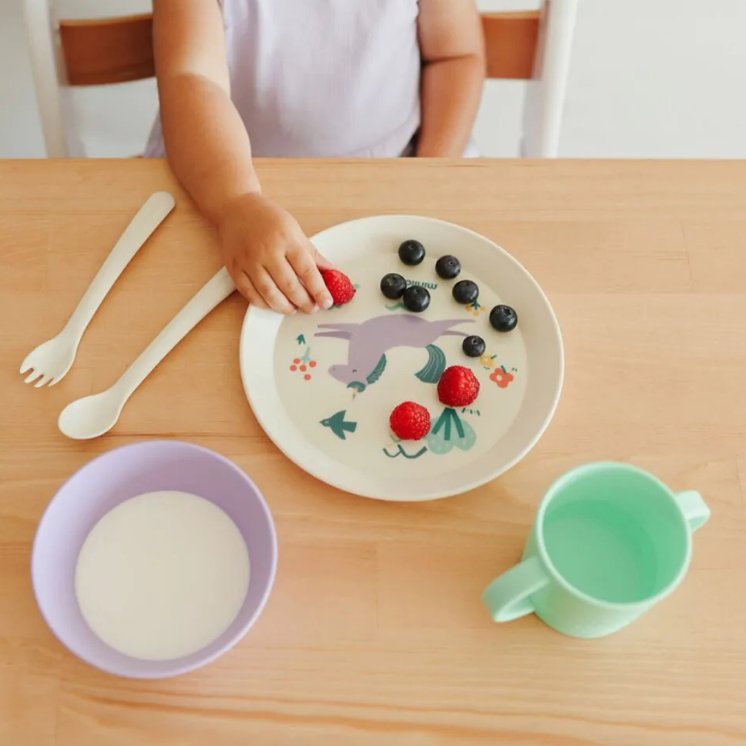 Conjunto infantil de refeição com prato, taça, copo e talheres sobre mesa de madeira clara.
