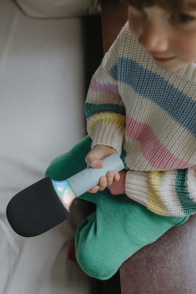 Criança a segurar microfone cinza claro com cápsula preta usando camisola de riscas coloridas e calças verdes