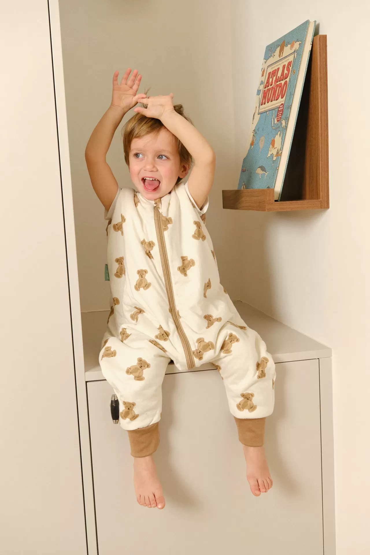 Criança com macacão de bebé creme e ursinhos castanhos sentada em móvel branco, perto de livro Atlas Mundo numa estante de madeira.