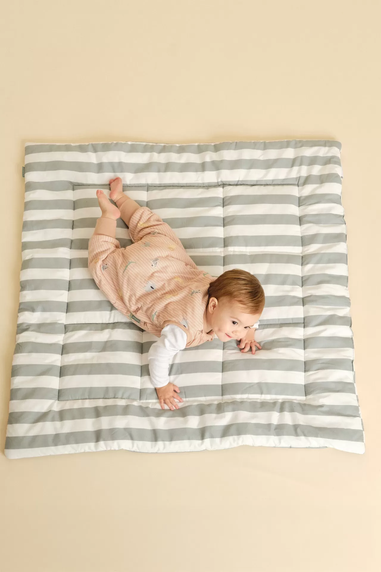 Bebé em macacão rosa claro num tapete acolchoado às riscas cinza e branco.