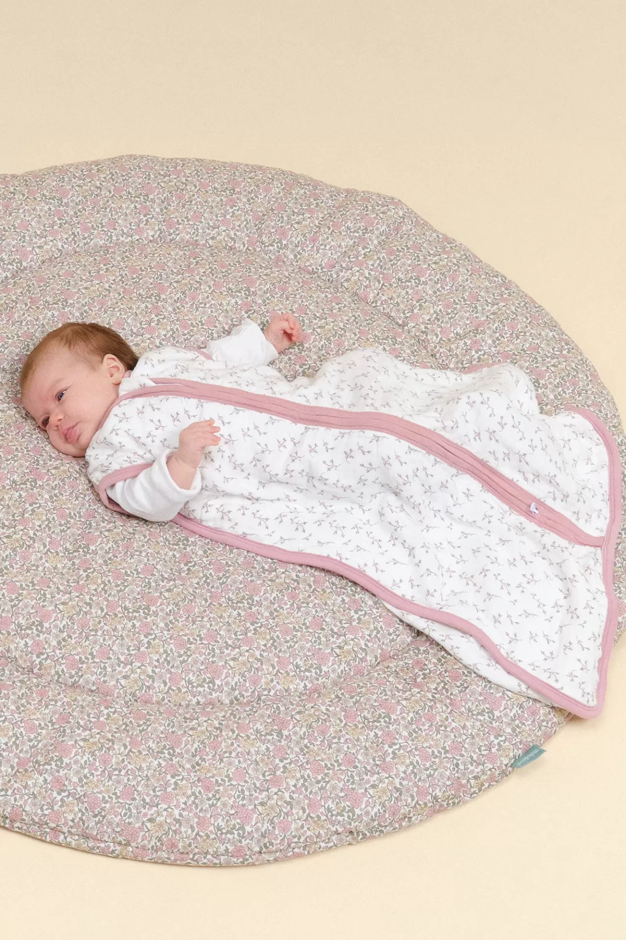 Bebé numa cama redonda macia com padrão floral rosa e saco de dormir branco com flores
