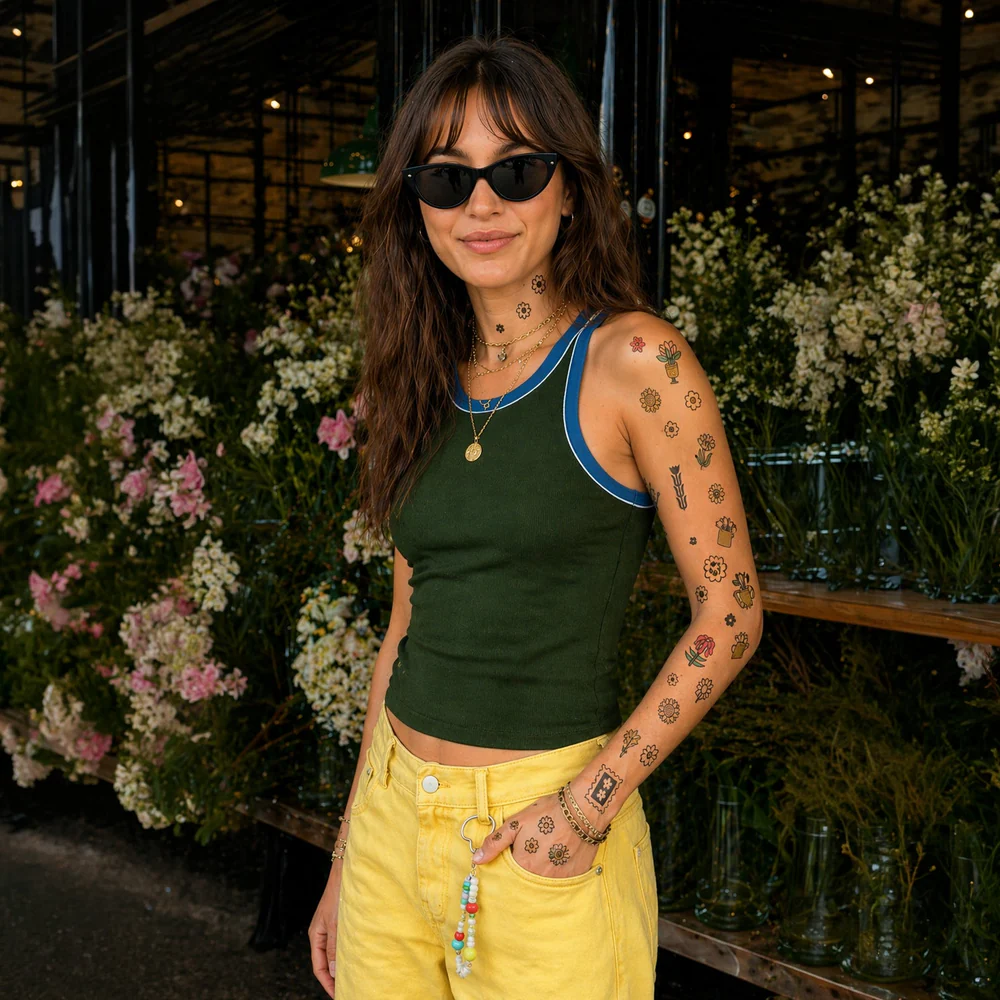 Pessoa com tatuagens temporárias florais, regata verde, calças amarelas e óculos escuros em ambiente florido
