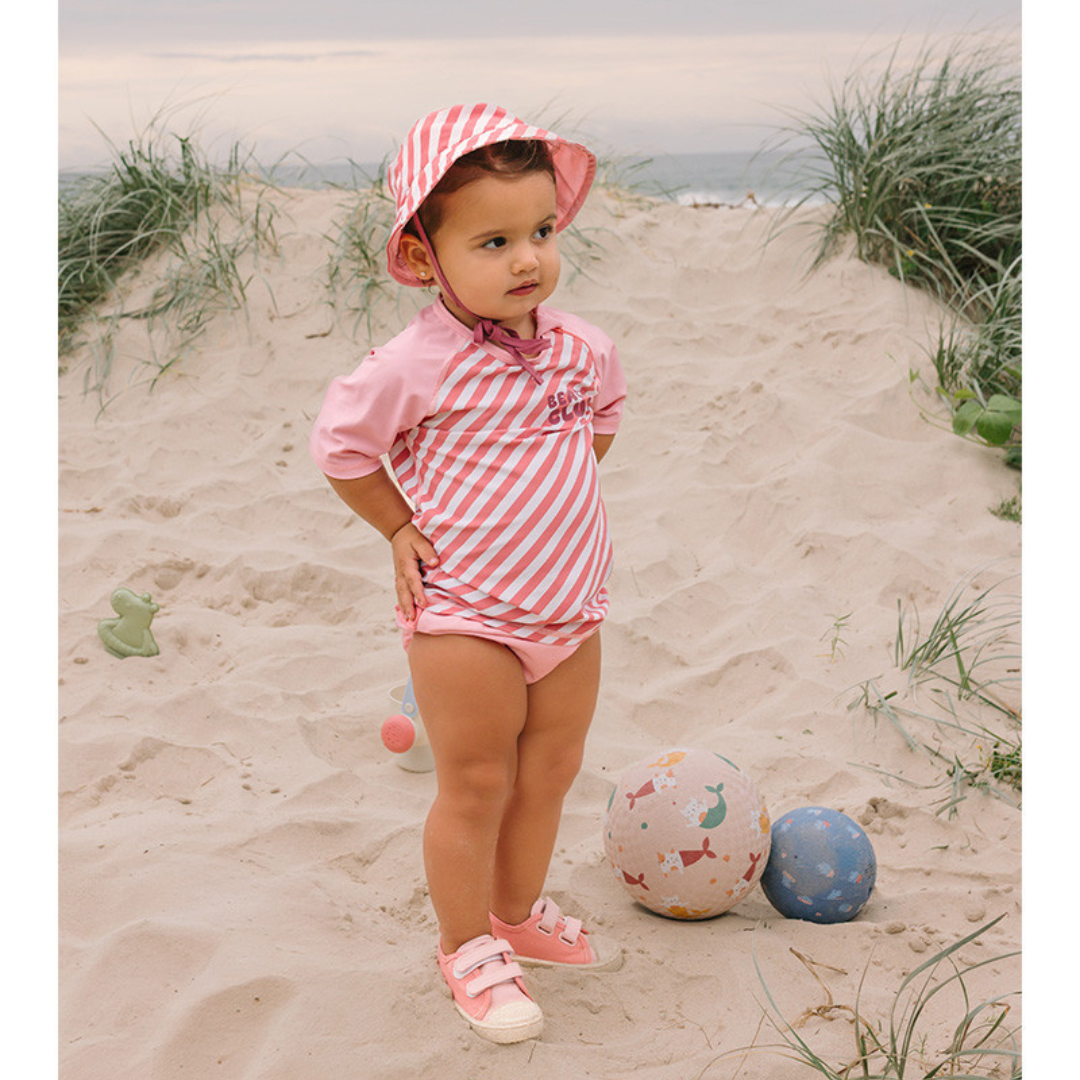 Criança com roupa de praia rosa em areia junto a bola colorida e vegetação