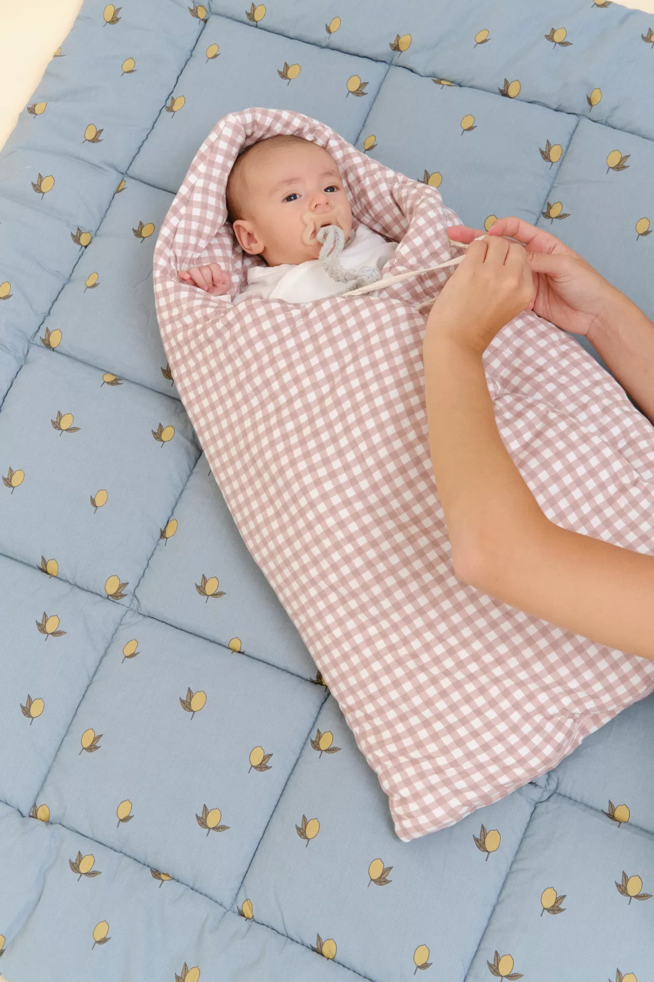 Bebé enrolado em saco xadrez bege e branco sobre tapete azul com limões amarelos.