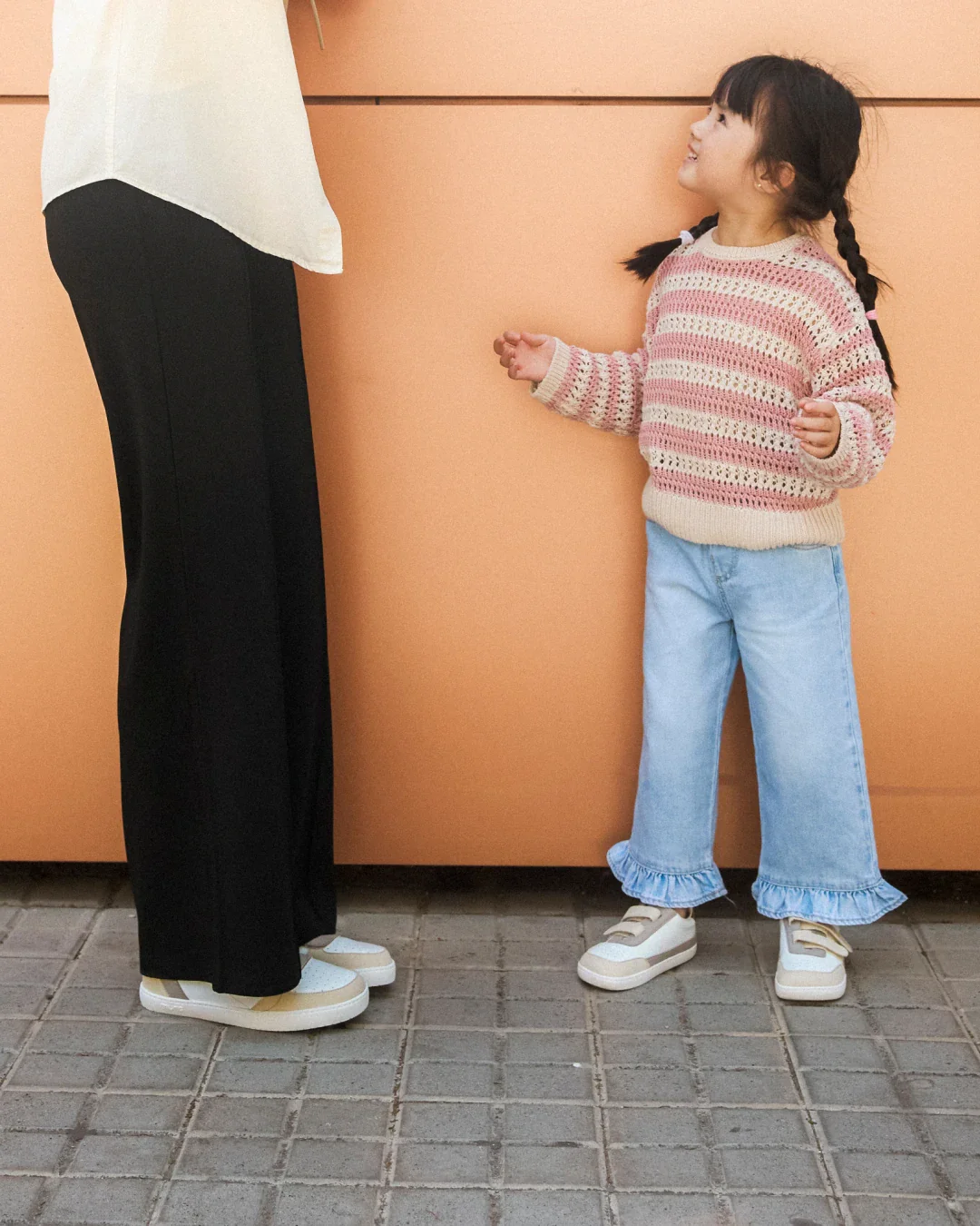 Criança com casaco de malha, calças jeans com folhos e sapatilhas junto a adulto com calças pretas e sapatilhas