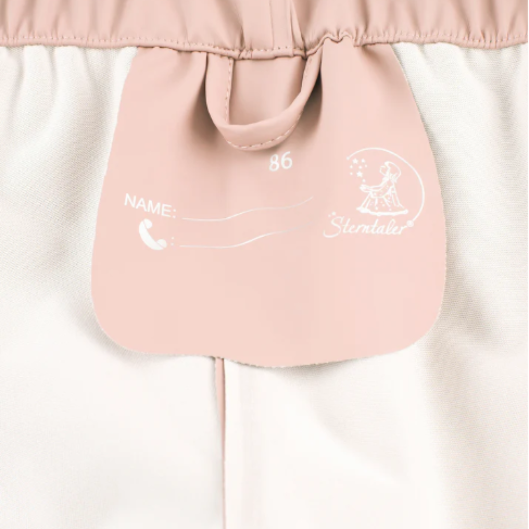 Etiqueta cor de rosa clara numa peça de roupa infantil branca com texto em branco.