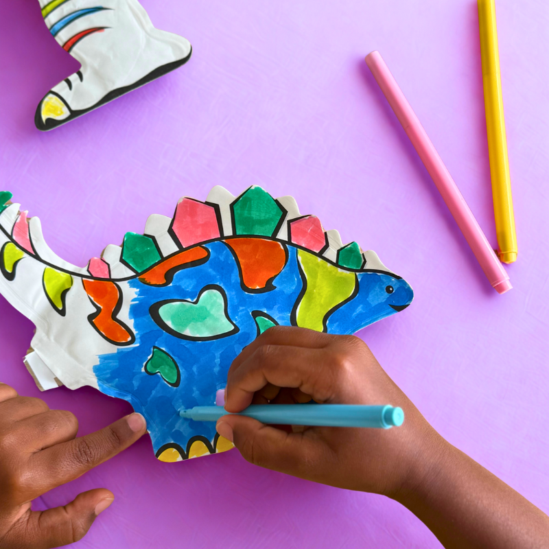 Mãos a colorir um dinossauro de papel com canetas coloridas sobre fundo lilás