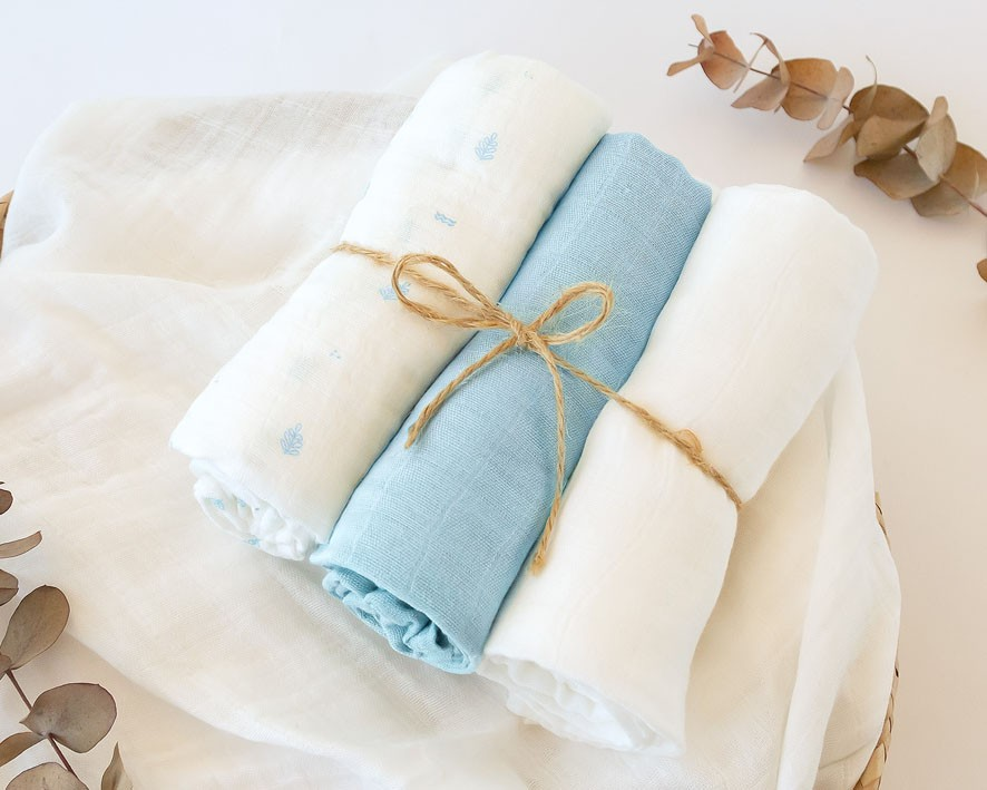 Três panos de tecido enrolados, dois brancos e um azul, com corda a amarrar e folhas secas ao lado