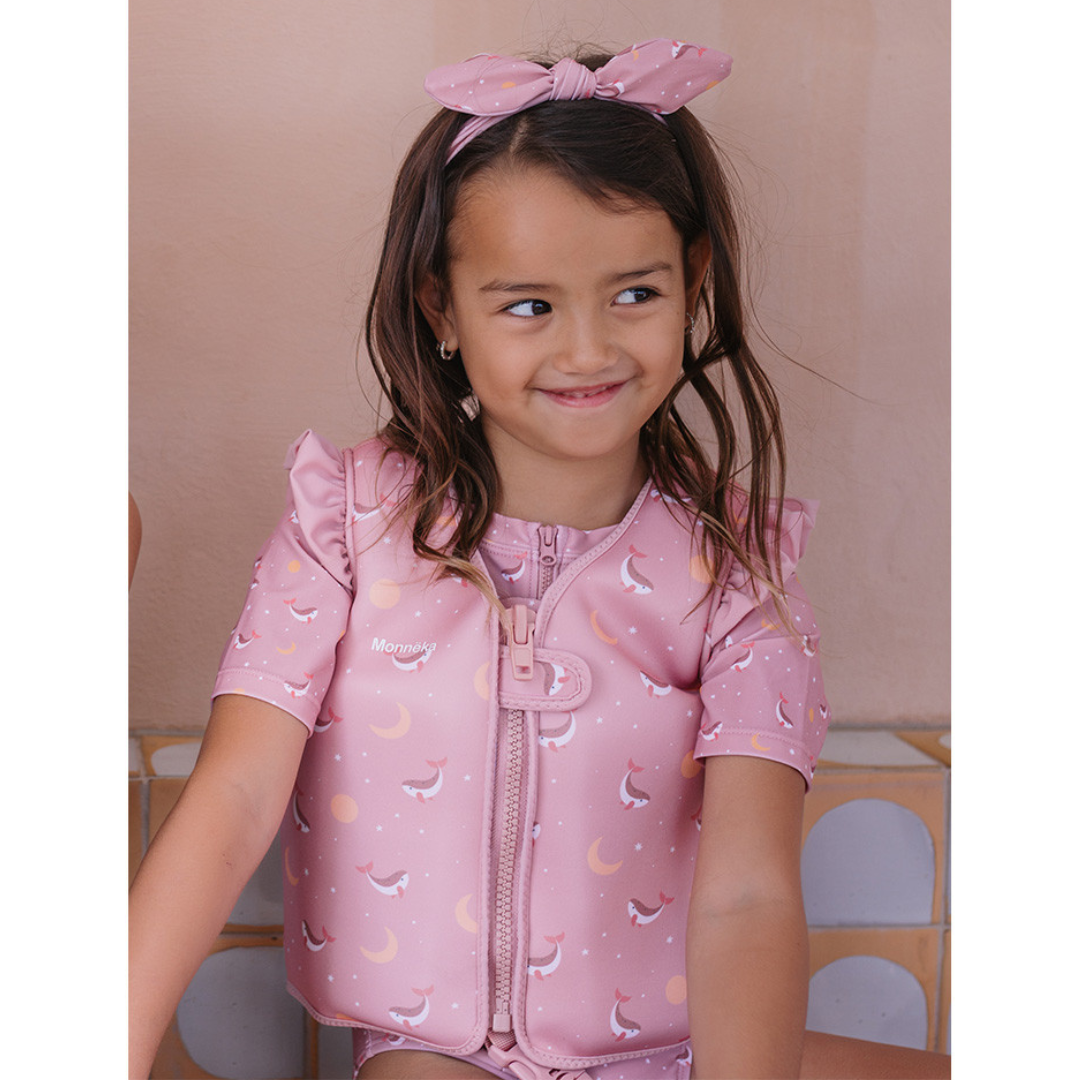 Menina com camisola de neoprene cor-de-rosa estampada e laço cor-de-rosa na cabeça sentada num banco de azulejos.