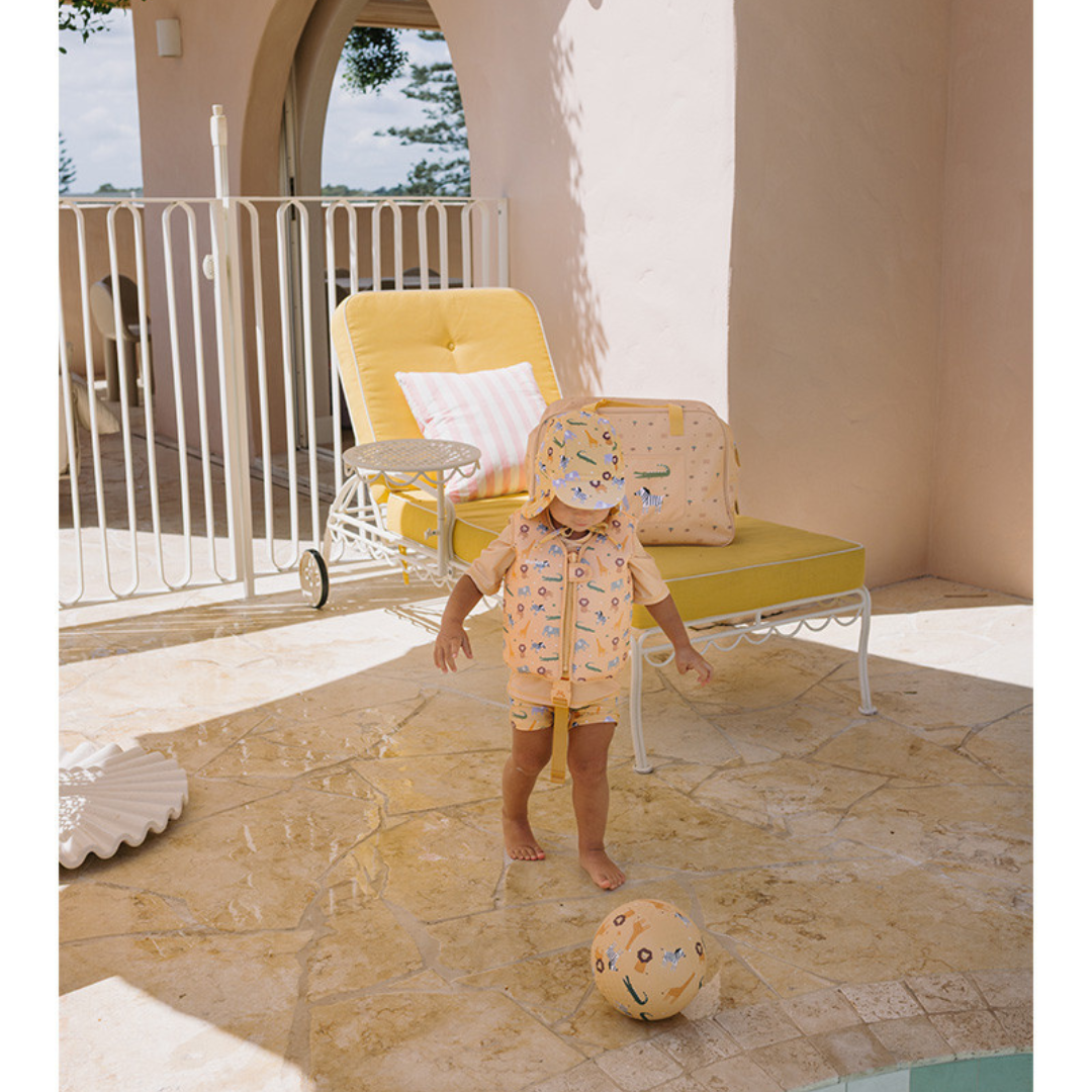 Criança vestida com roupa cor de pêssego com estampados a brincar com bola à beira de uma piscina ao lado de espreguiçadeira amarela.