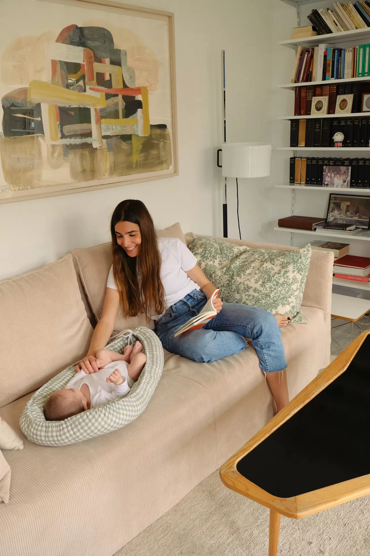 Sofá bege com almofada verde floral, bebê em poltrona xadrez e mesa de centro de madeira clara
