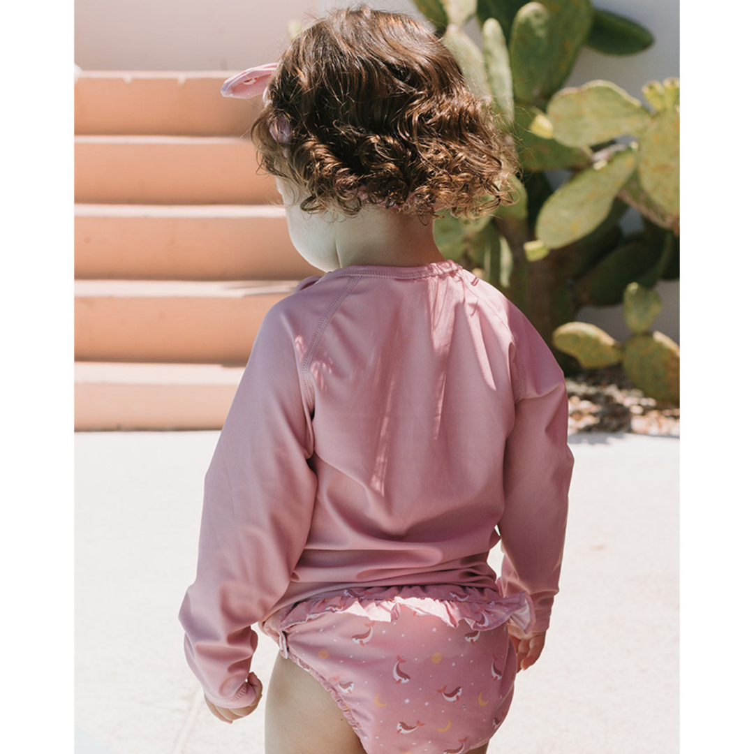Criança com roupa de banho rosa com padrão de luas e estrelas perto de escadas exteriores cor de salmão e cactos