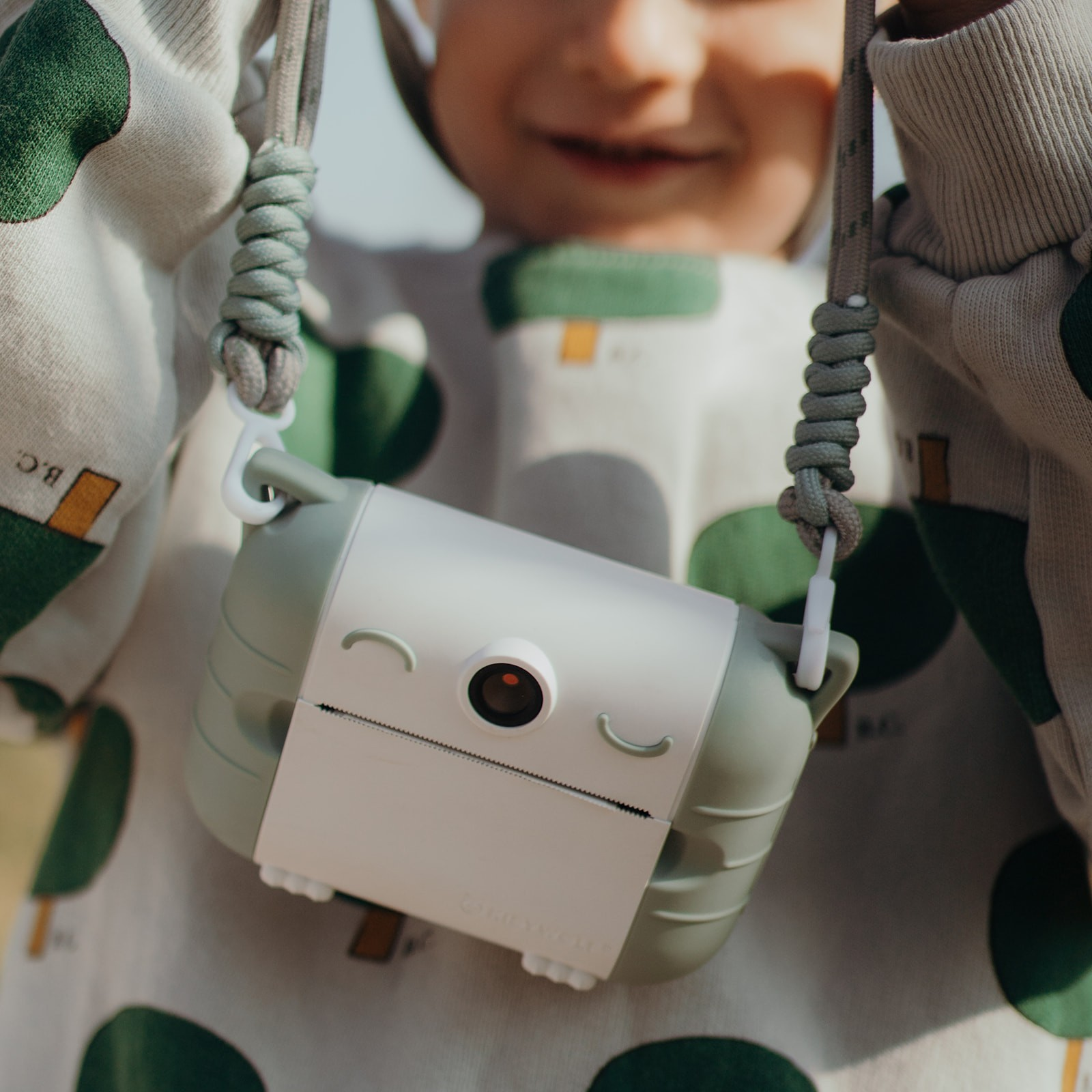Máquina fotográfica branca com rosto sorridente pendurada no pescoço de criança com camisa branca e verde