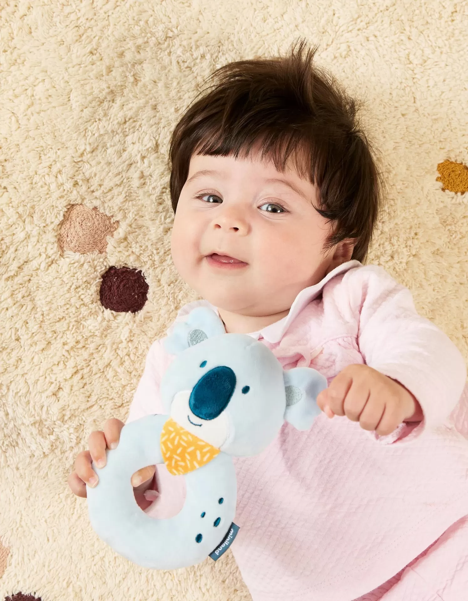 Bebé a segurar brinquedo koala de peluche azul claro, sobre tapete bege com círculos.