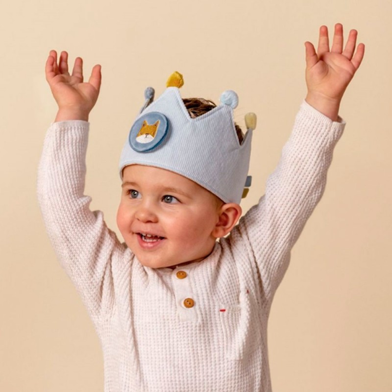 Criança com coroa azul e camisola bege com botões em fundo bege