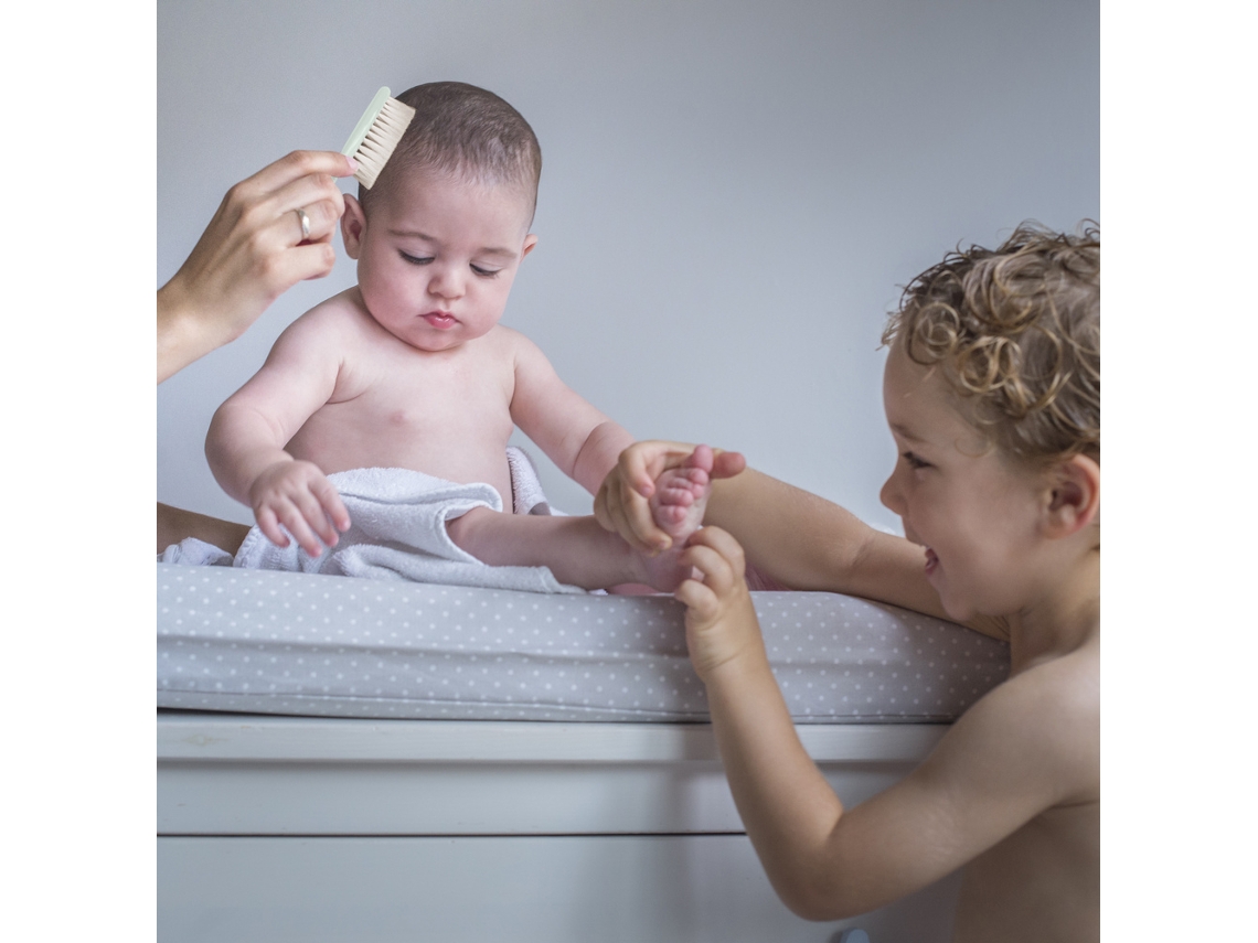 Bebé sentado numa superfície almofadada cinza com bolinhas brancas com criança a brincar ao lado e mão a segurar um pente branco.