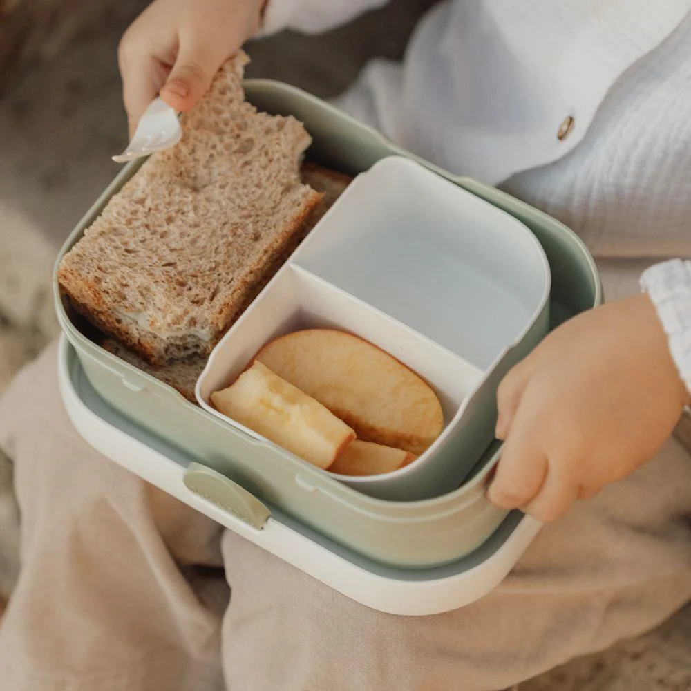 Marmita com sandes, maçã e queijo segurada por criança com camisa branca