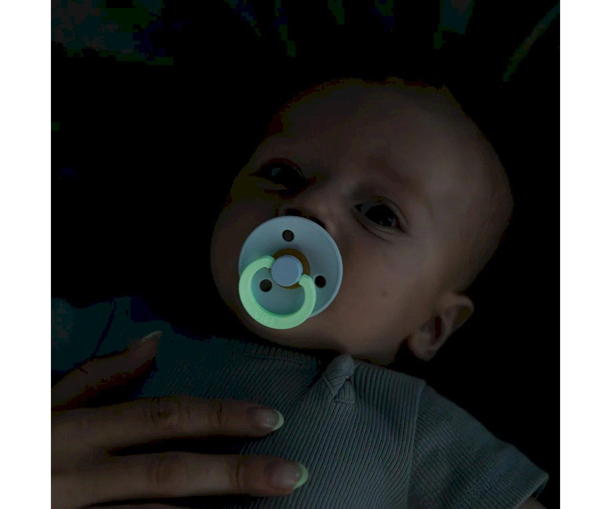 Bebé a usar chupeta luminosa verde fosforescente com camisola cinza clara e mão de adulto no peito