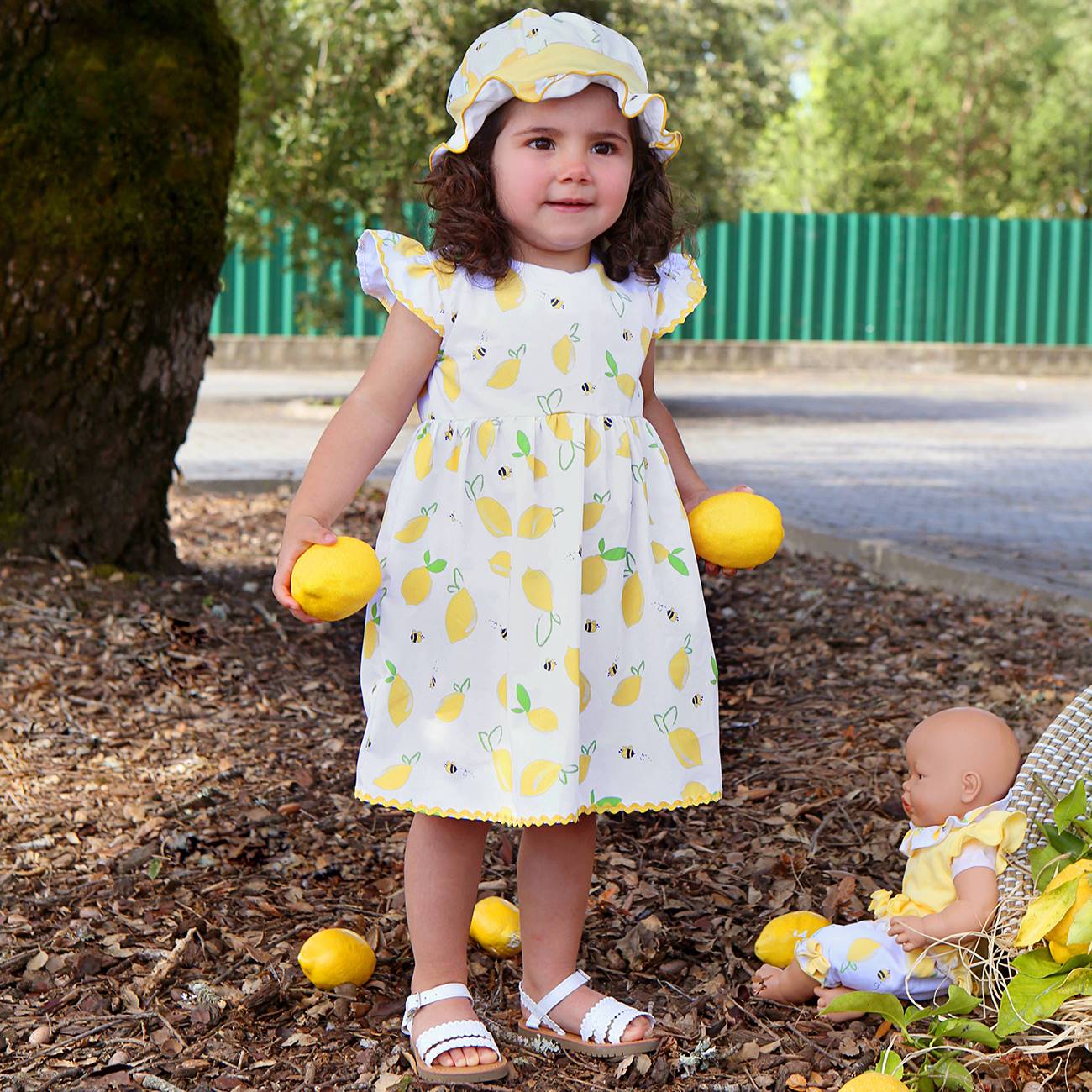 Menina com vestido e chapéu branco com estampado de limões amarelos segurando limões em ambiente exterior
