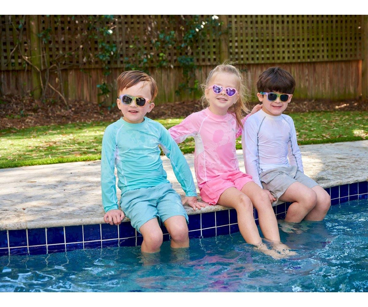 Três crianças na beira de piscina com óculos de sol e roupas de banho de manga longa