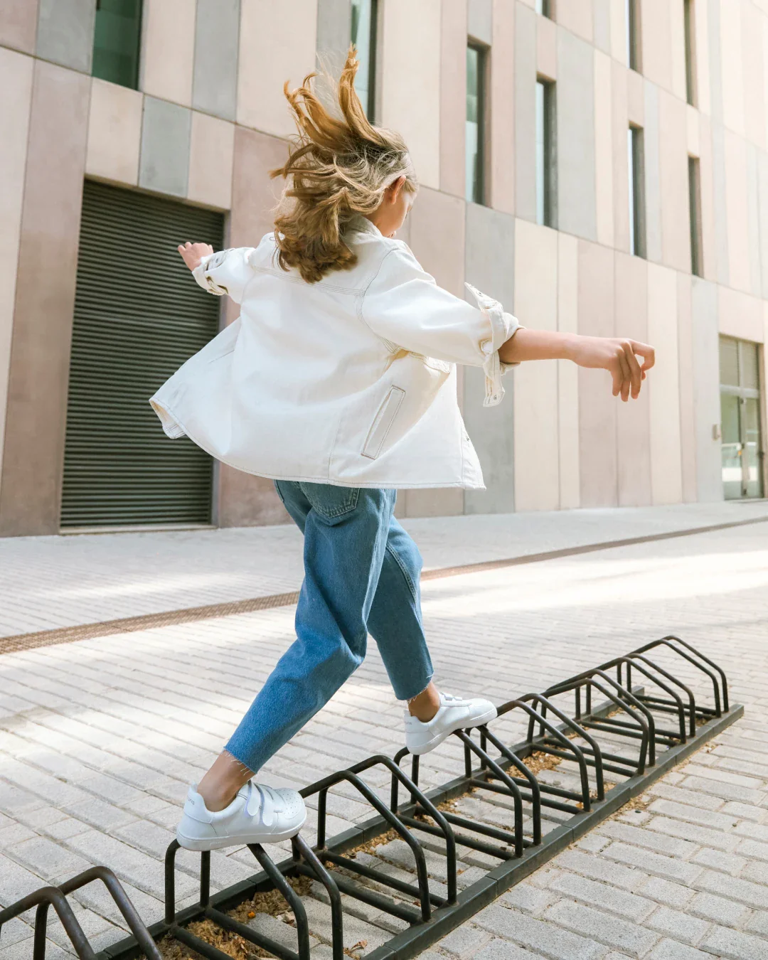 Pessoa a equilibrar-se com casaco branco, calças de ganga e ténis brancos numa rua com edifícios modernos