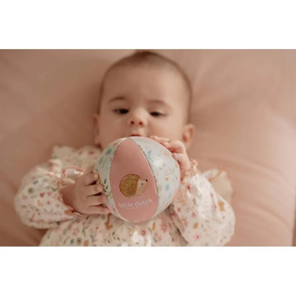 Bebé segurando bola de brinquedo macia com padrão floral e painel rosa com ouriço.