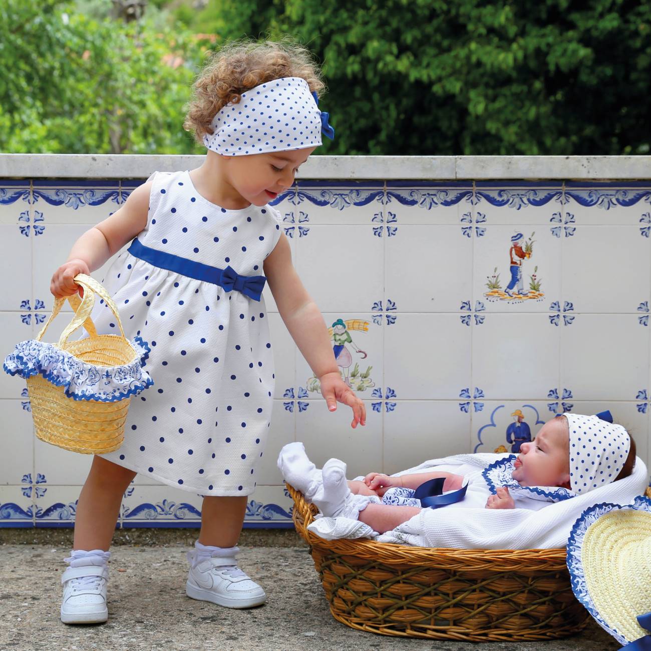 Criança vestindo vestido branco com bolinhas azuis e faixa azul, ao lado de um bebé deitado em cesto de vime com roupa azul e branca, frente a uma parede de azulejos decorativos