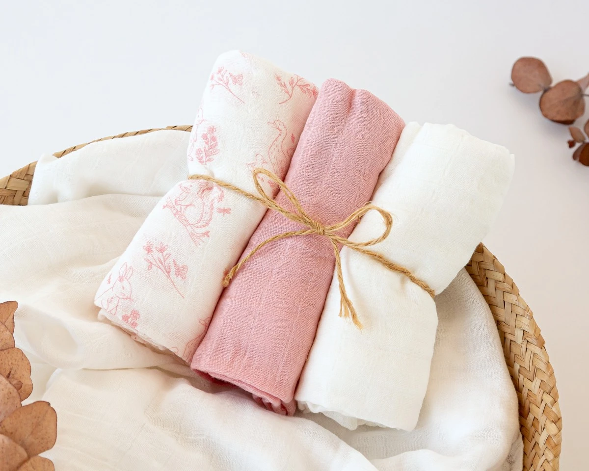 Três panos de tecido macio, um estampado rosa, outro rosa liso e outro branco, enrolados e amarrados com corda numa cesta de vime.