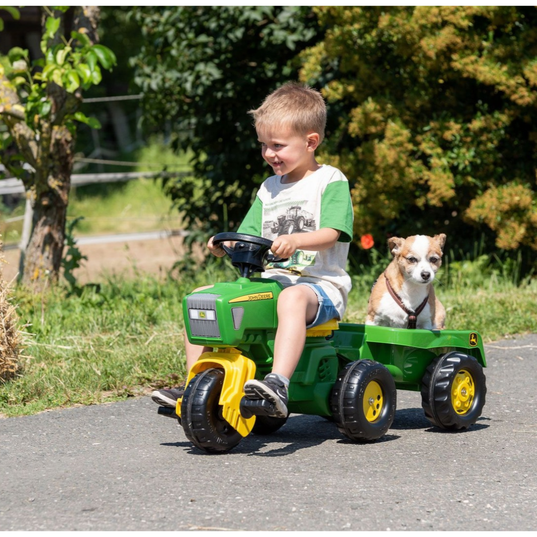 Criança num tractor de brincar verde e amarelo John Deere com um cão sentado no atrelado ao lado, em ambiente exterior