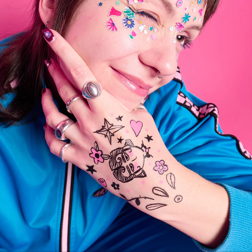 Mão com tatuagens temporárias coloridas e anéis, rosto com autocolantes florais, fundo rosa.