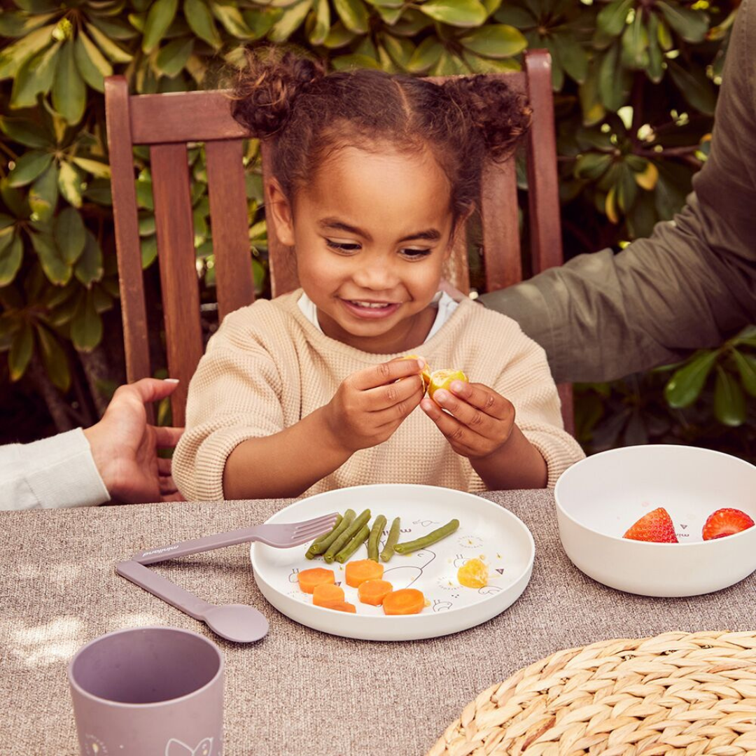 Criança a comer tangerina com prato branco e talheres lilás numa mesa com toalha cinzenta