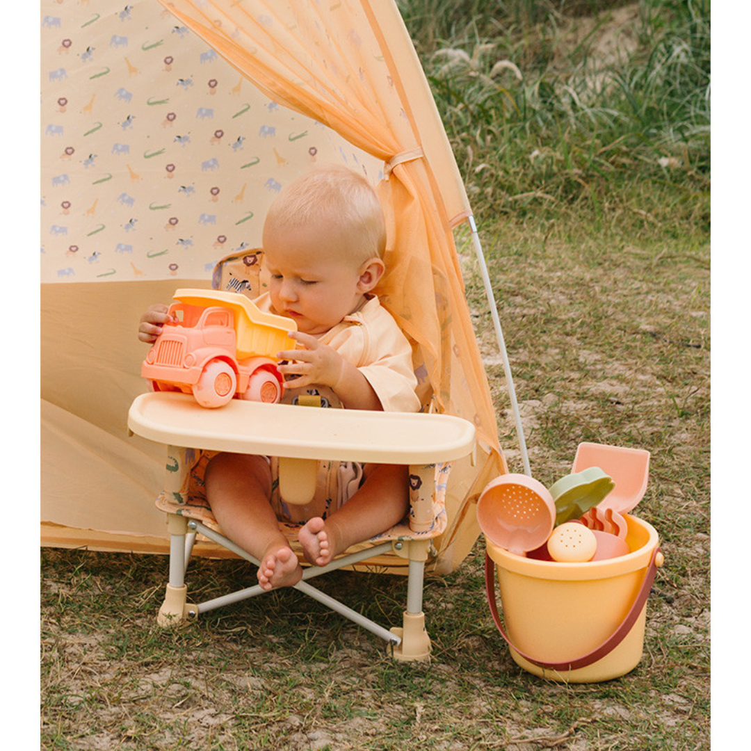 Bebé sentado a brincar com um camião de brinquedo numa cadeira junto a um balde amarelo com brinquedos, num espaço com relva e uma tenda decorada com animais.