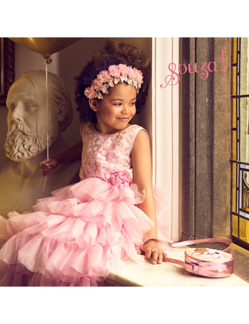 Menina de vestido rosa com coroa de flores e balão dourado sentada numa janela ao lado de um busto de mármore.