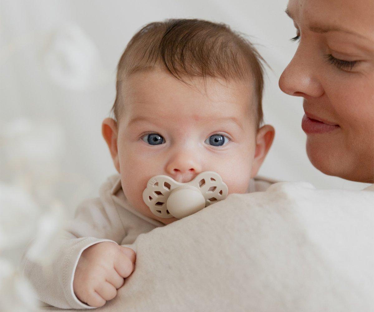 Bebé com chupeta branca e adulto segurando, fundo claro desfocado