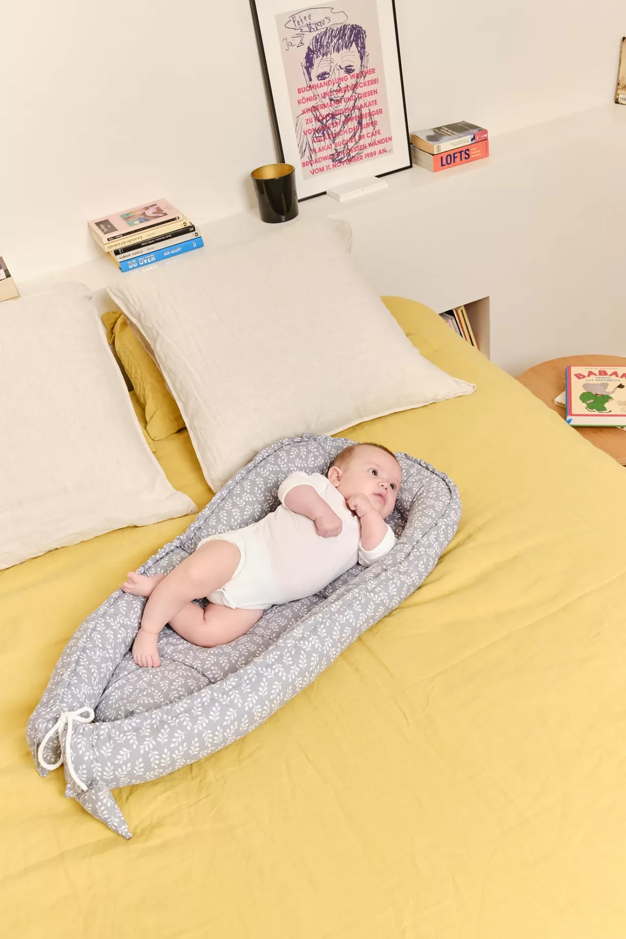 Almofada para bebé cinzenta com padrão branco sobre cama amarela com almofadas e bebé de body branco deitado