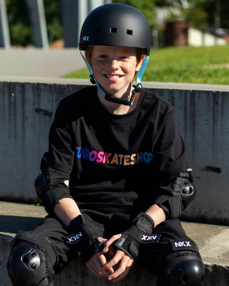 Menino com capacete preto, equipamento de proteção preto e camisola preta com letras coloridas sentado ao ar livre.