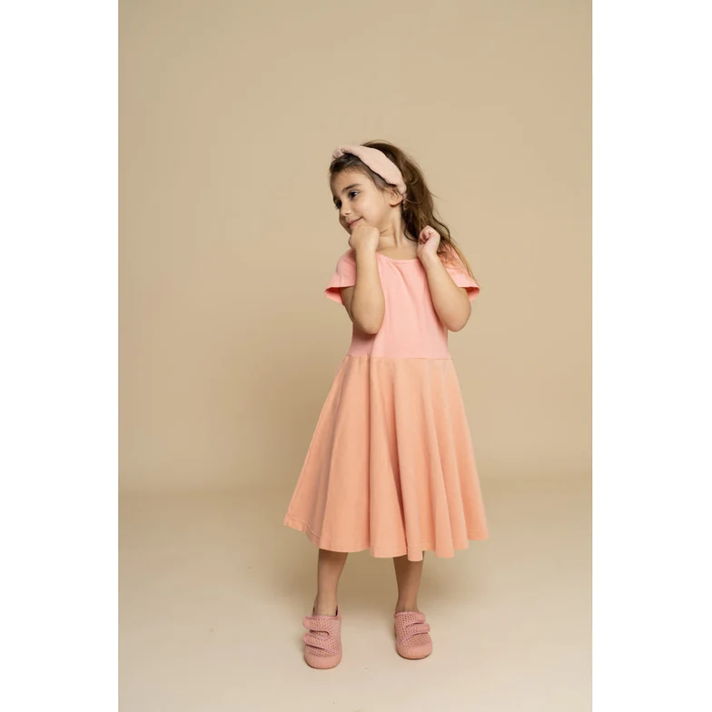 Menina com vestido rosa claro, faixa de cabeça e sapatos rosas num fundo bege.