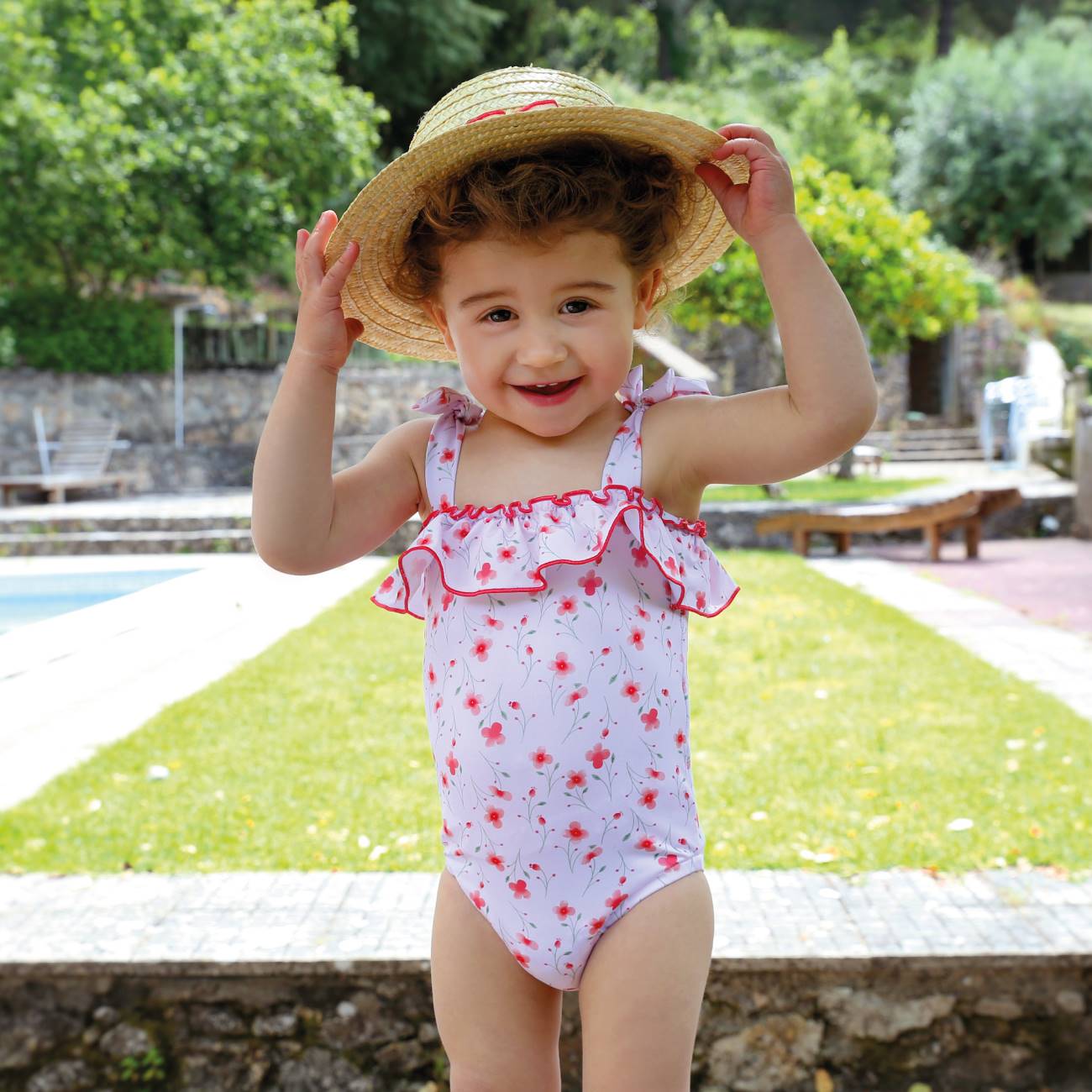 Menina com fato de banho branco com flores vermelhas e chapéu de palha num jardim com piscina
