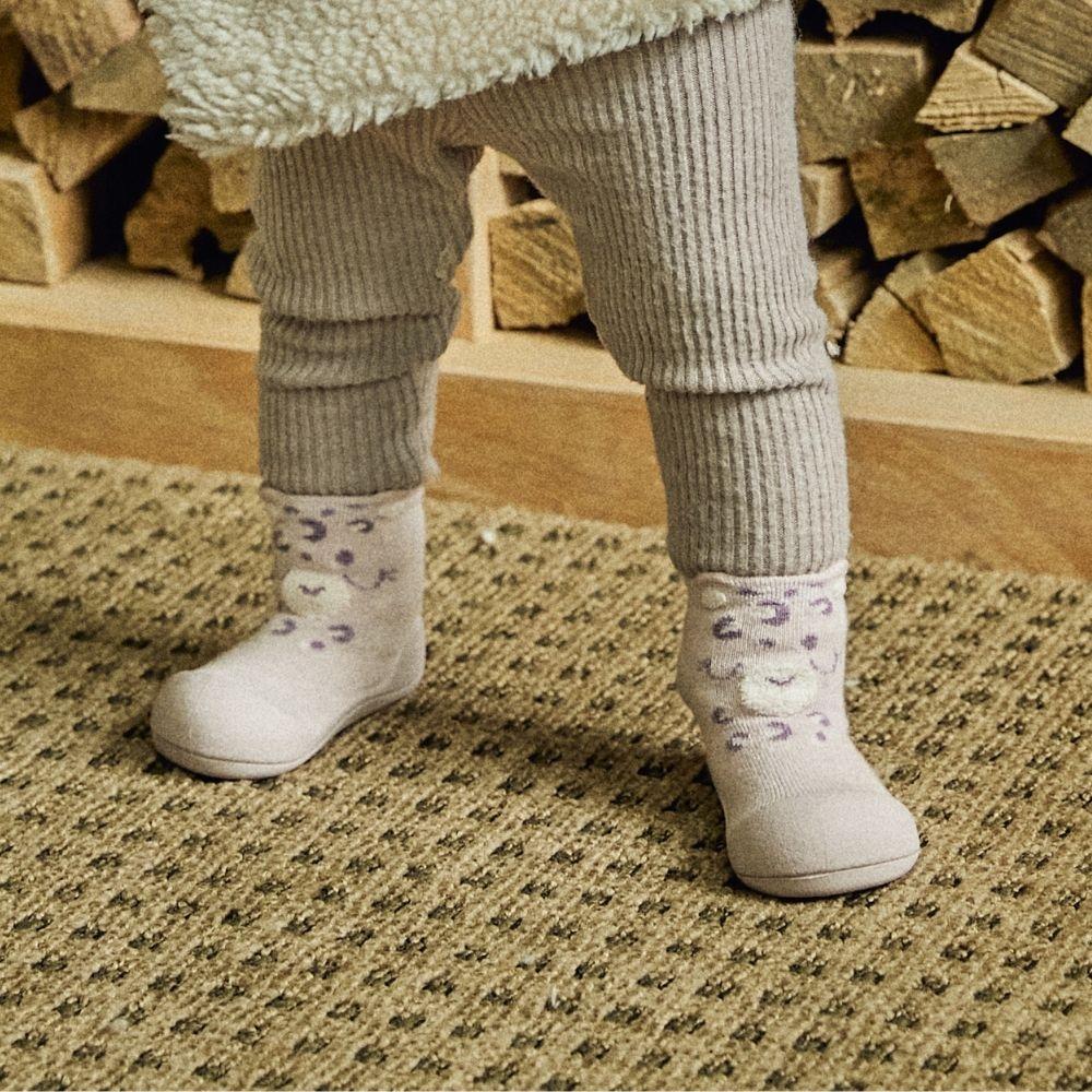 Bebé com calças cinzentas e sapatinhos brancos com padrão roxo e rosto sorridente