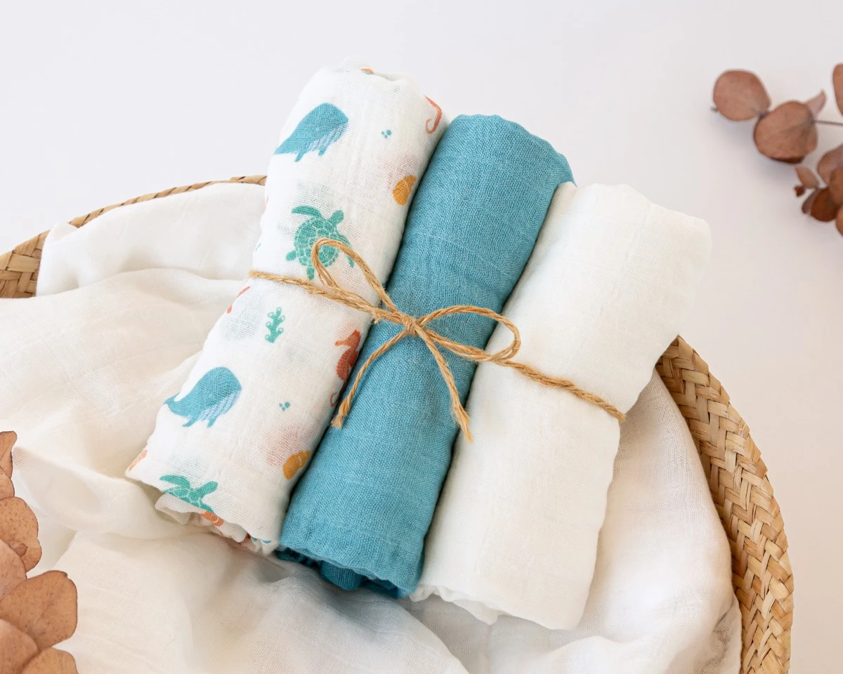 Três toalhas enroladas, uma com padrão de animais marinhos, uma azul e uma branca, amarradas com cordão de juta num cesto de verga.