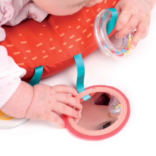 Espelho redondo cor-de-rosa com fita azul e almofada cor-de-laranja com padrão, mãos de bebé e brinquedo colorido