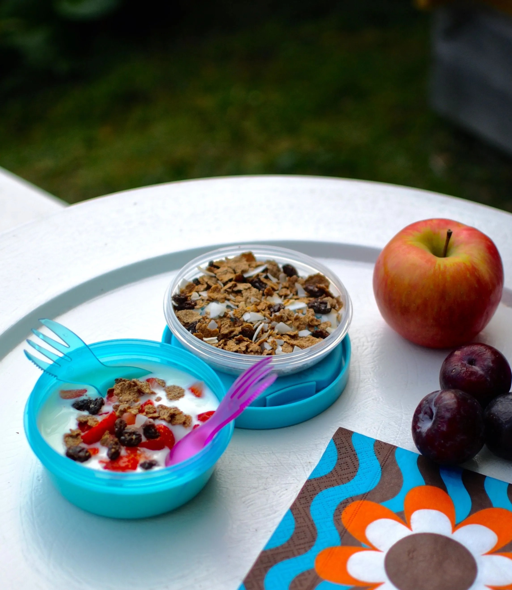Recipientes azuis com iogurte e granola, maçã, ameixas e guardanapo colorido numa mesa branca redonda ao ar livre