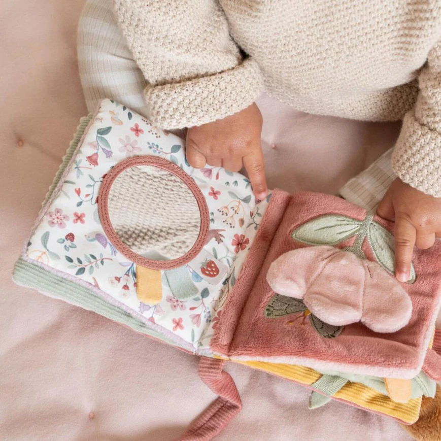 Livro infantil de tecido com padrões florais, espelho e flor em relevo, mãos de criança a folhear.