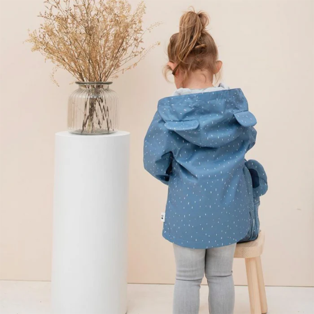 Criança com casaco azul e capuz, sentada de costas num banco de madeira ao lado de vaso com flores secas