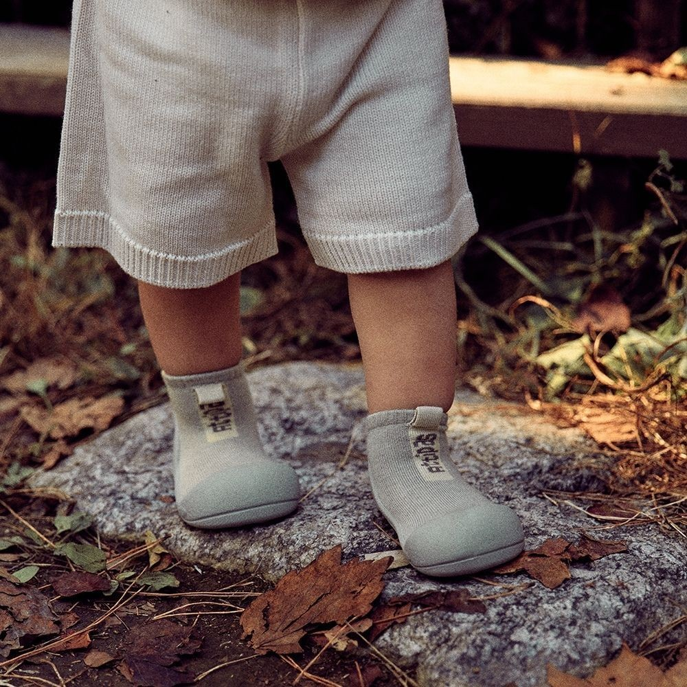 Criança com calças de malha bege e calçado infantil cinzento com sola flexível ao ar livre